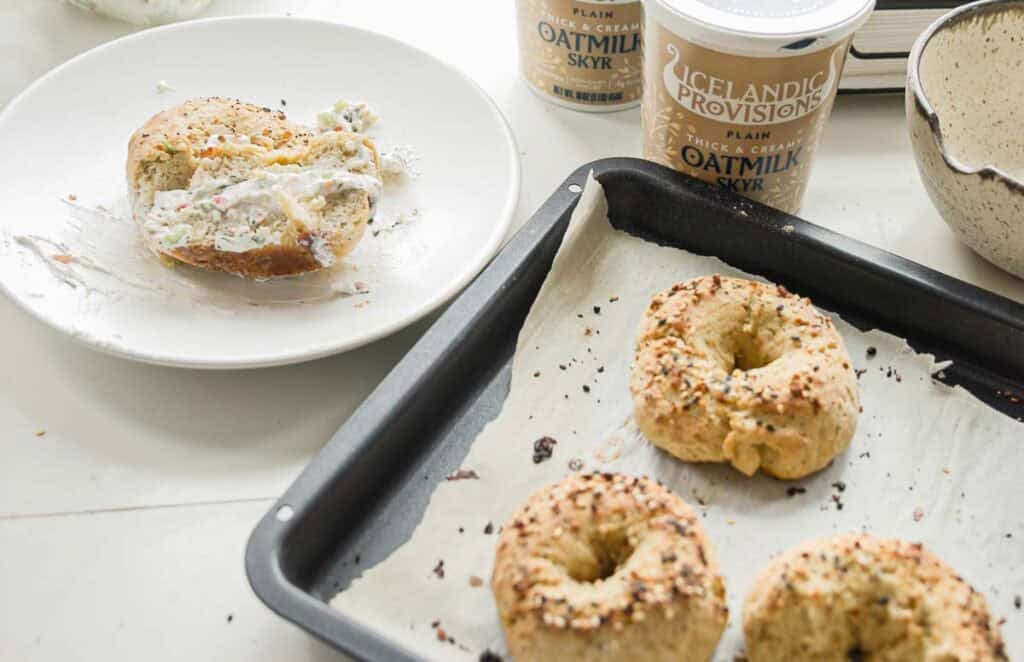 Freshly baked bagels on a baking sheet with one bagel cut and spread with a creamy topping on a plate. Containers of Icelandic Provisions plain oat milk skyr are in the background on a light-colored surface.